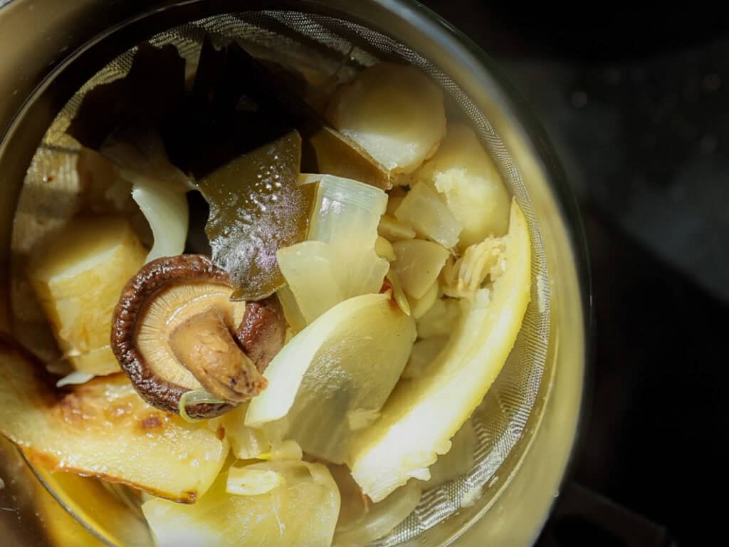 A close-up of a metal strainer containing various cooked vegetables, including sliced carrot, onions, and a whole mushroom, with visible steam and a warm light illuminating the ingredients.